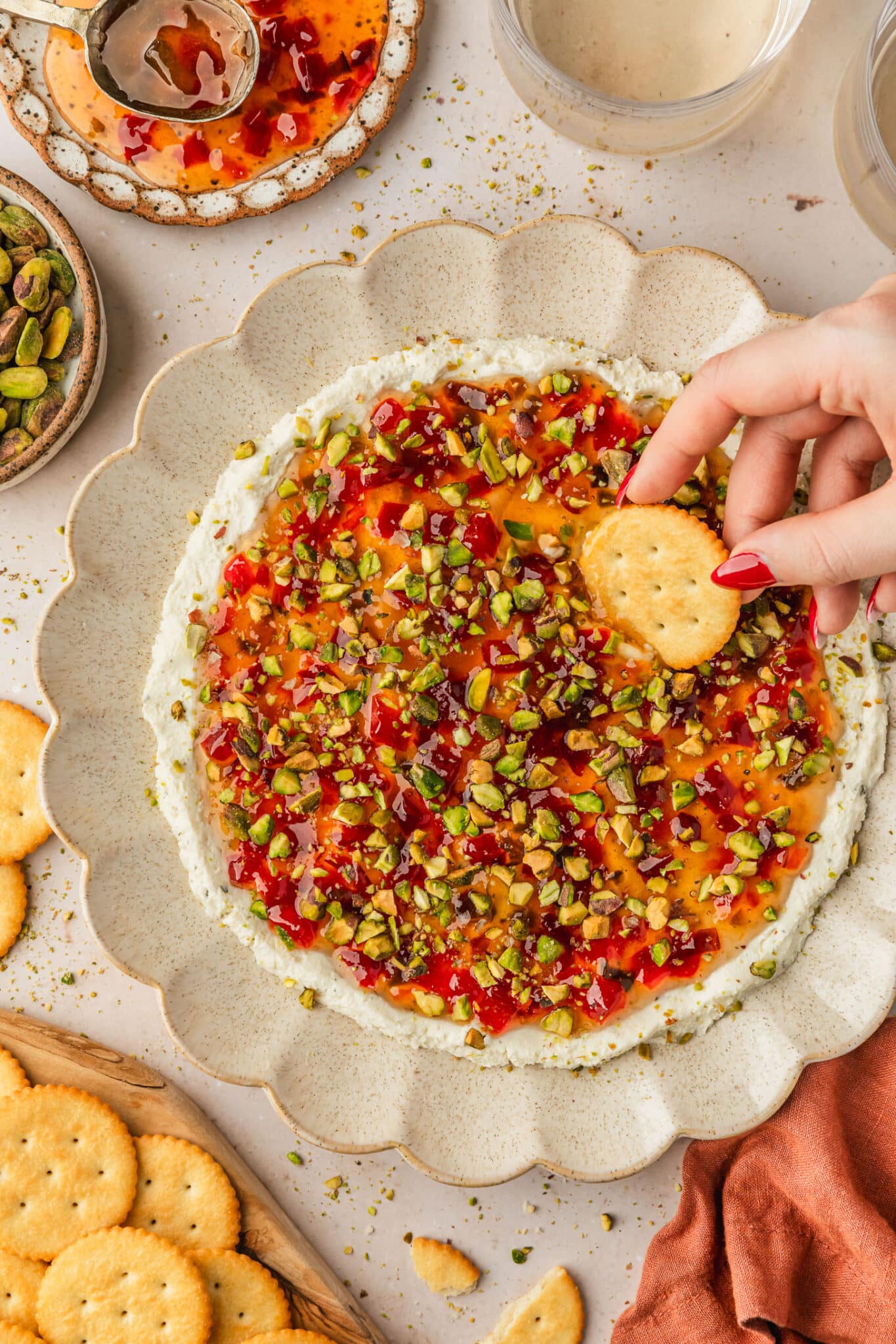 A hand dipping a cracker into a tan platter of boursin dip with pepper jelly and pistachios on a tan counter next to glasses of white wine, an orange linen, and brown bowls of jelly, pistachios, and crackers.