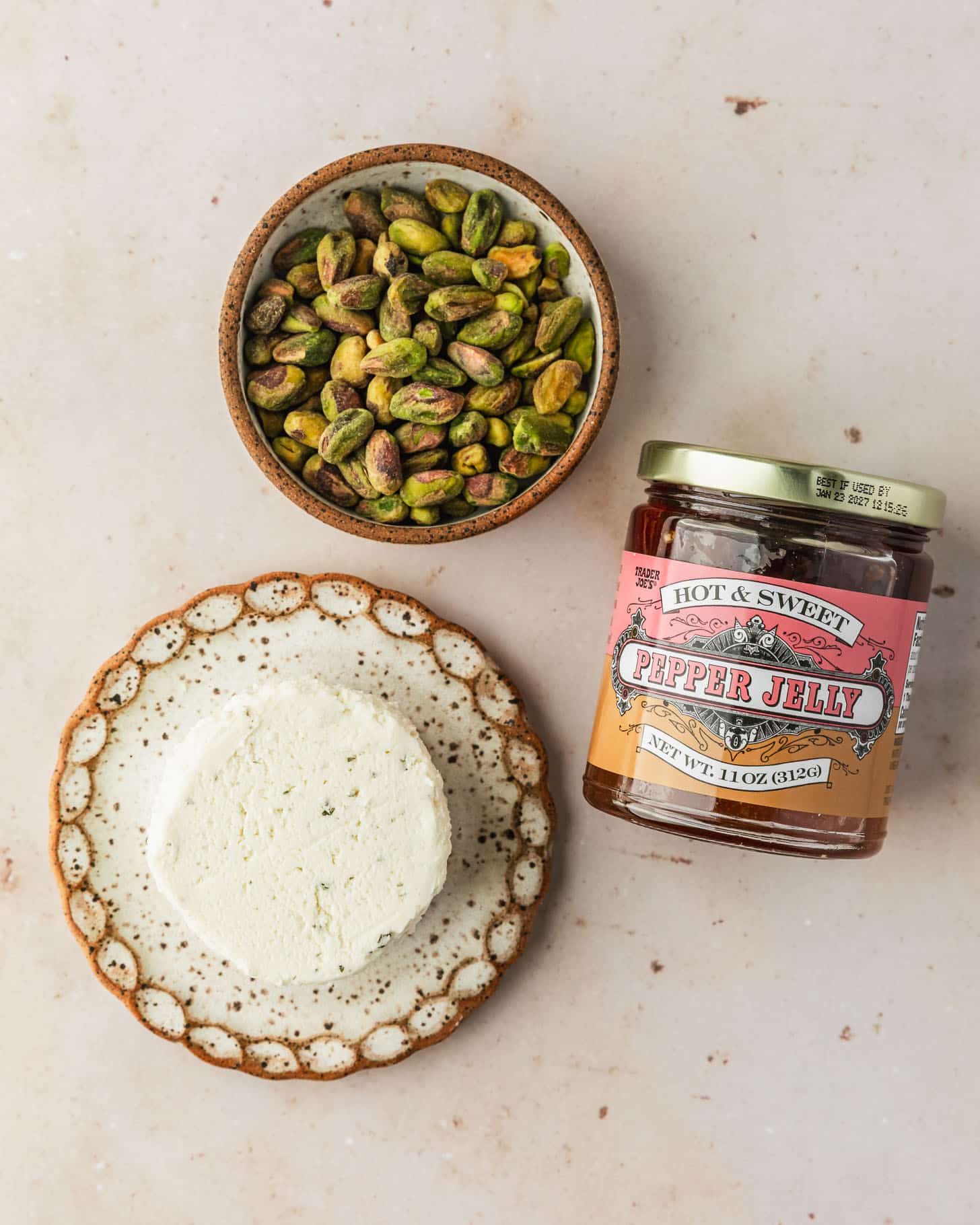 A white plate of cheese, a brown bowl of pistachios, and a jar of pepper jelly on a tan marble counter.