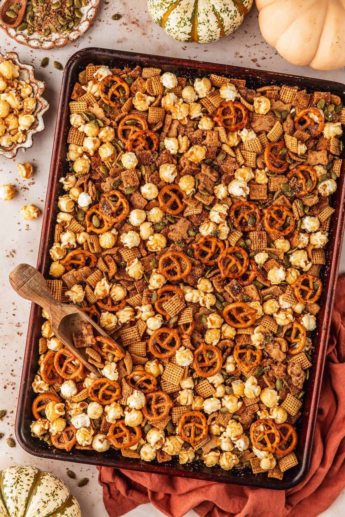 A sheet pan with autumn snack mix on a tan counter next to an orange linen, pumpkins, and brown bowls of caramel popcorn and pepitas.
