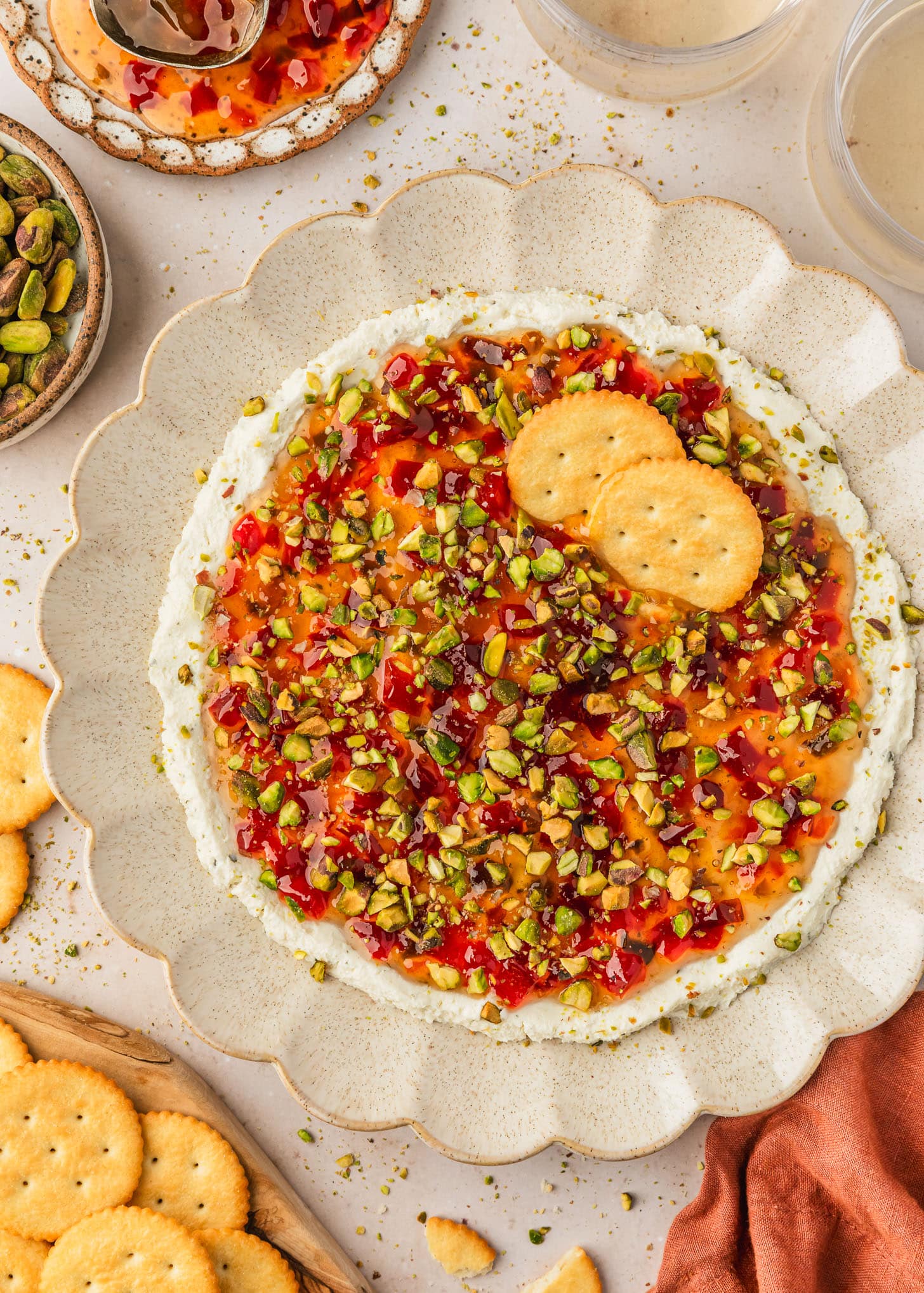 A beige plate of 3 ingredient boursin dip with pepper jelly and pistachios on a tan counter next to glasses of white wine, an orange linen, and brown bowls of crackers, pistachios, and jelly.