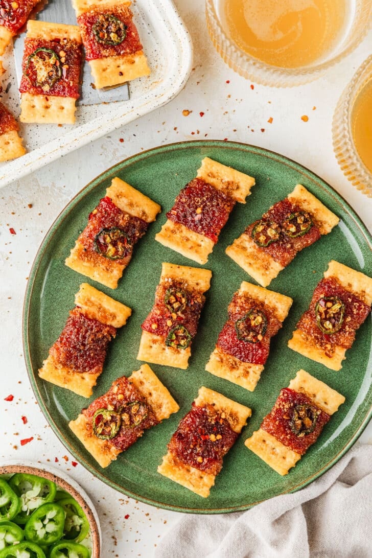 A green plate of candied bacon crackers on a tan counter next to a beige linen, glasses of beer, white pan of crackers, and white bowl of jalapenos.