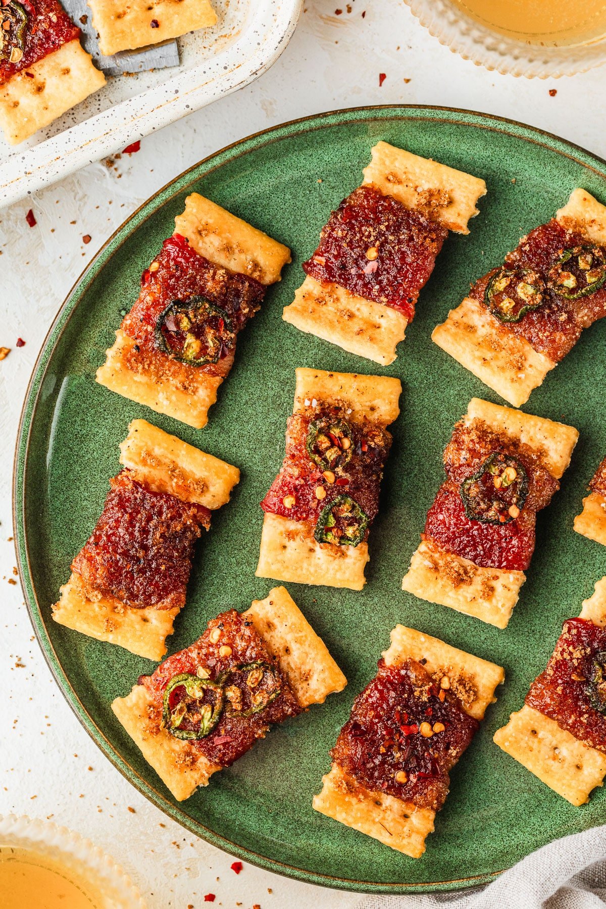 A green platter of spicy candied bacon wrapped crackers on a white table next to beer, a white pan of crackers, and a white bowl of peppers.
