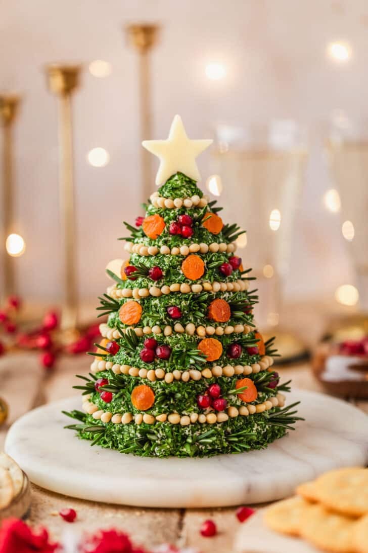 A marble board with a Christmas tree cheese ball on a wood table next to glasses of champagne, a red linen, gold candlesticks, and brown bowls of crackers.