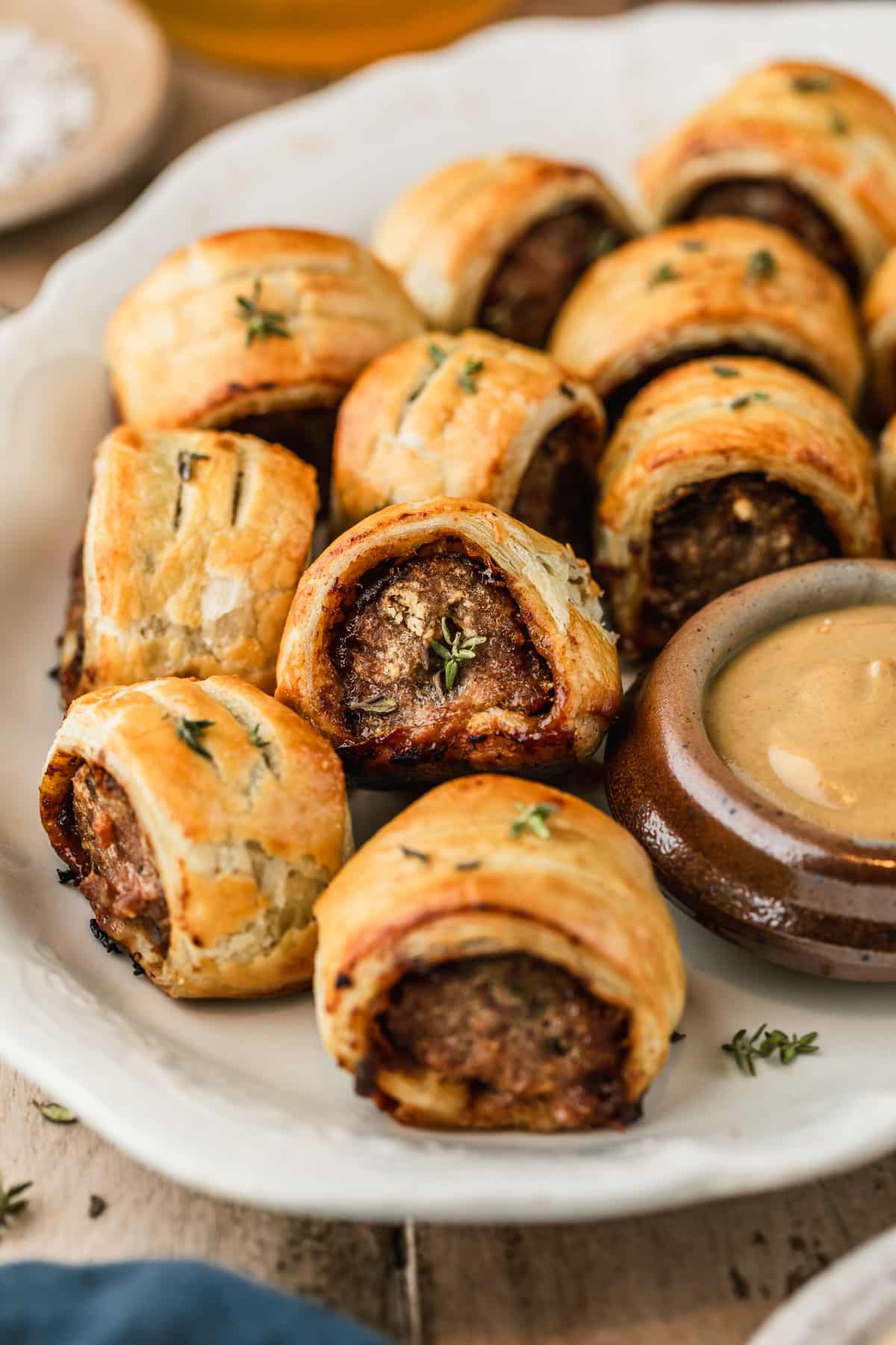 Pork and apple sausage rolls and a brown bowl of honey mustard on a vintage white platter next to a glass of beer and blue linen with a wood background.