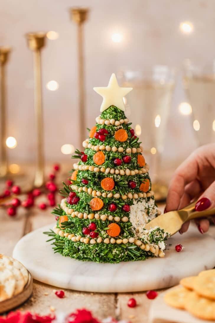 A hand using a gold knife to slice a Christmas tree cheese ball on a marble tray next to glasses of champagne, gold candlesticks, crackers, and a red linen with a wood background.