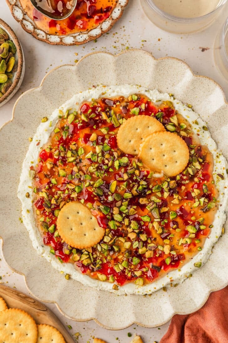 A tan platter of pepper jelly boursin dip with pistachios on a tan marble counter next to an orange linen, glasses of white wine, and brown bowls of jelly and pistachios.