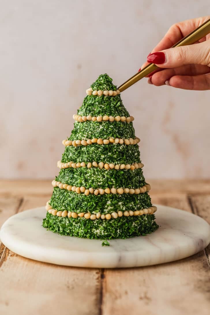 A hand using tweezers to decorate a Christmas tree cheese ball with pine nuts.