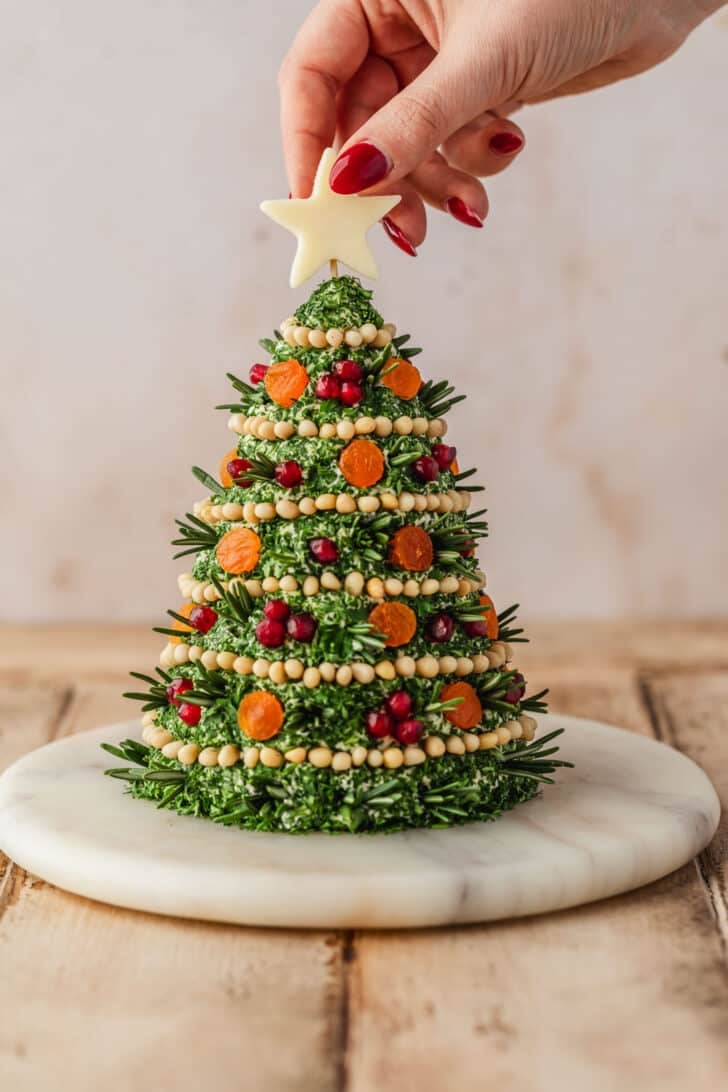 A hand placing a cheese star on a Christmas tree cheese ball with a tan backbround.