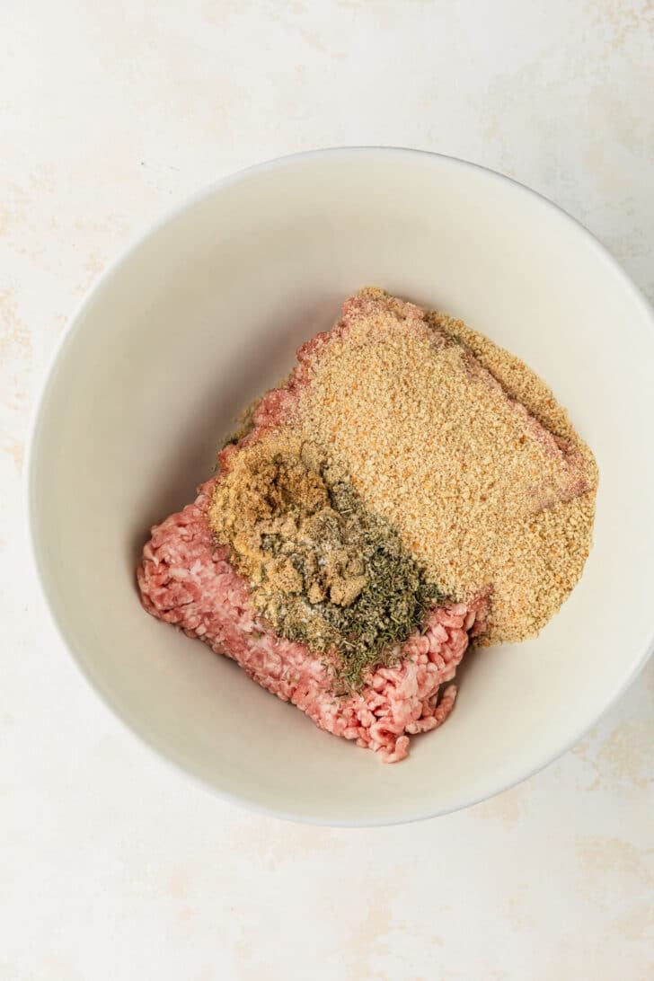 A white bowl of pork, spices, and breadcrumbs on a tan counter.