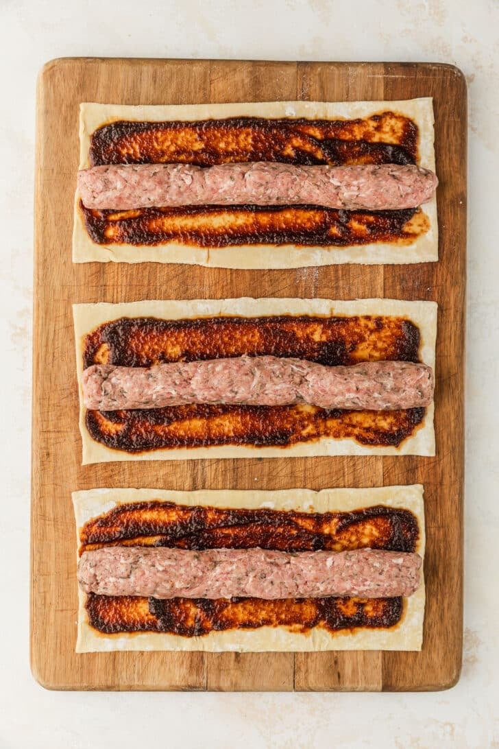 A wood board with three sheets of puff pastry topped with apple butter and sausage logs.