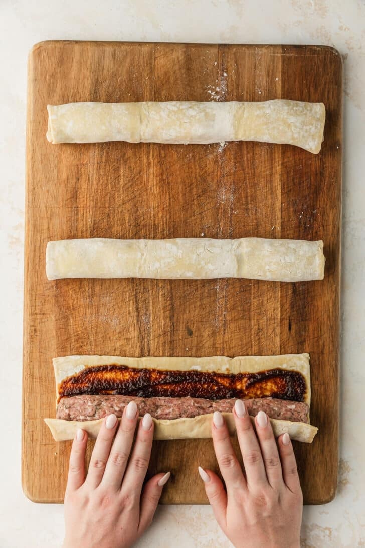 Hands rolling pork and apple sausage rolls on a wood board.