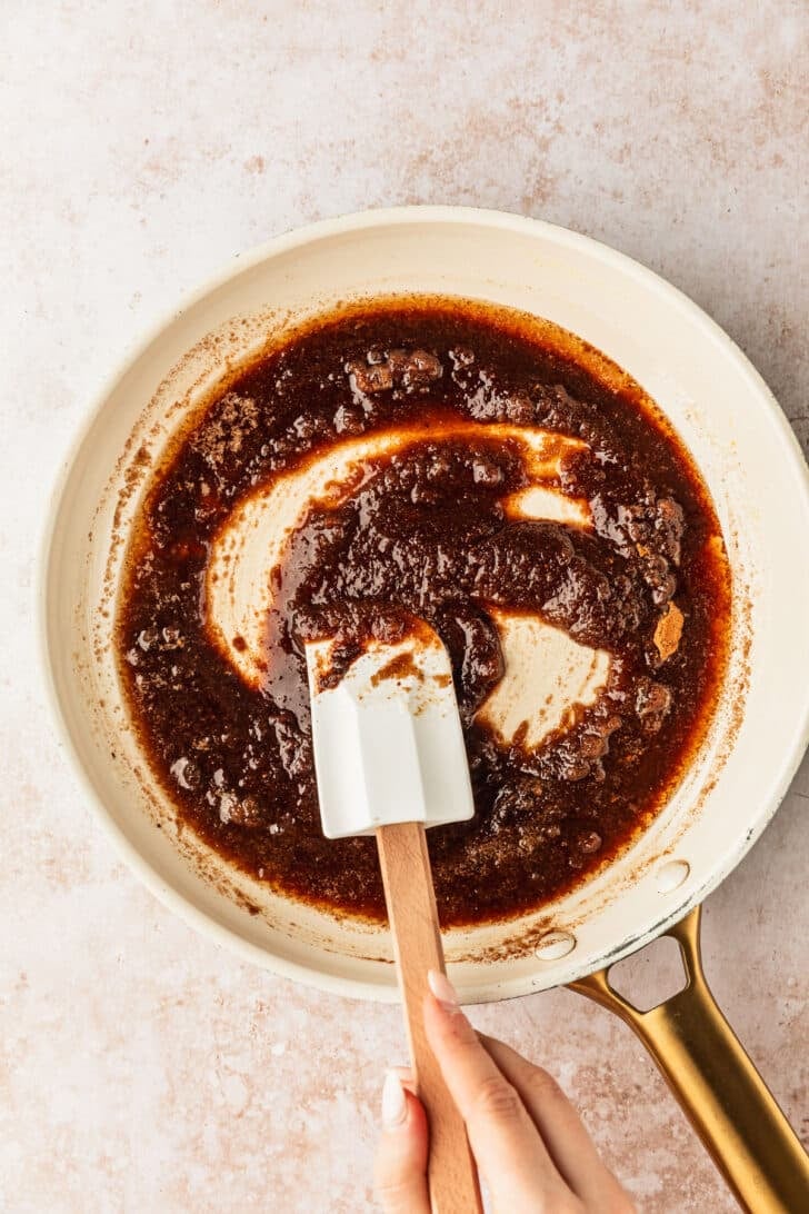 A hand stirring a white pan of brown sugar-butter mixture for autumn snack mix.