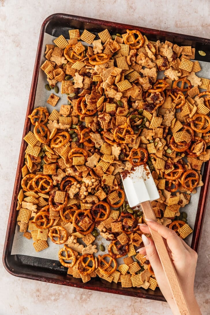 A hand using a rubber spatula to mix autumn snack mix on a pan.