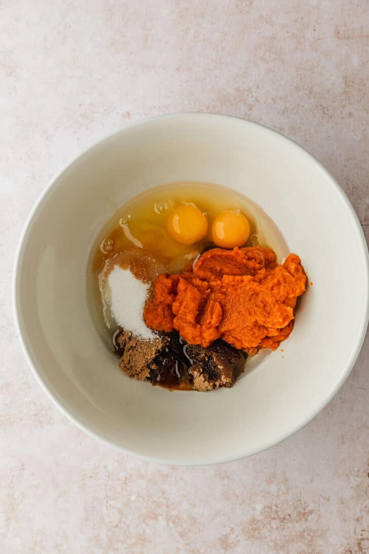 A white bowl of pumpkin, eggs, sugar, and oil on a tan counter.