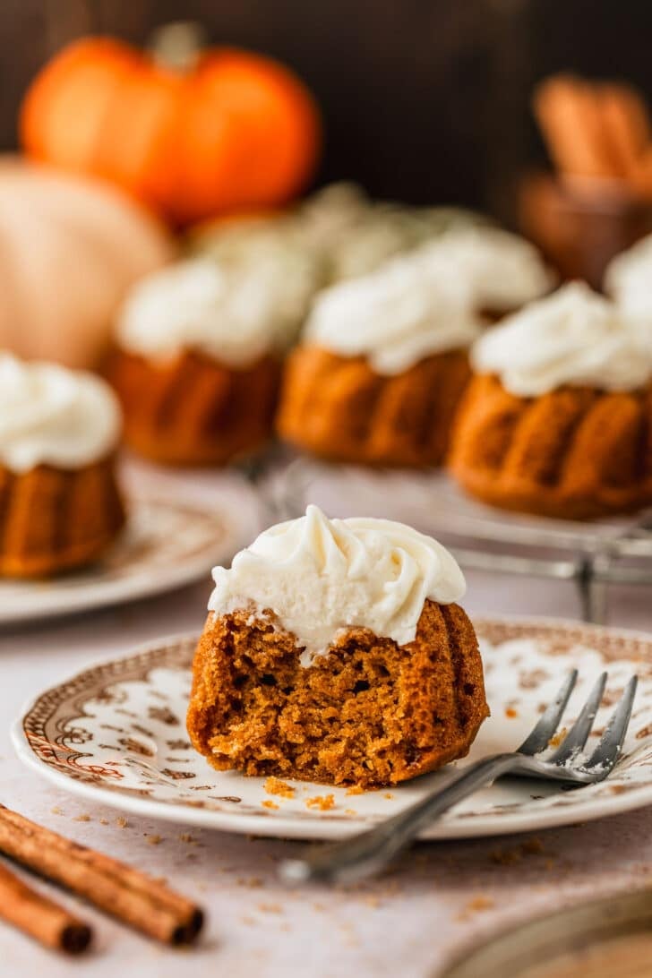 A brown plate with a cake on a tan counter next to pumpkins, cinnamon sticks, and a wire rack of pumpkin mini bundt cakes.