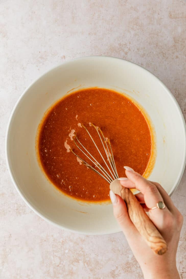 A hand using a whisk to mix pumpkin, eggs, oil, and sugars in a white bowl.