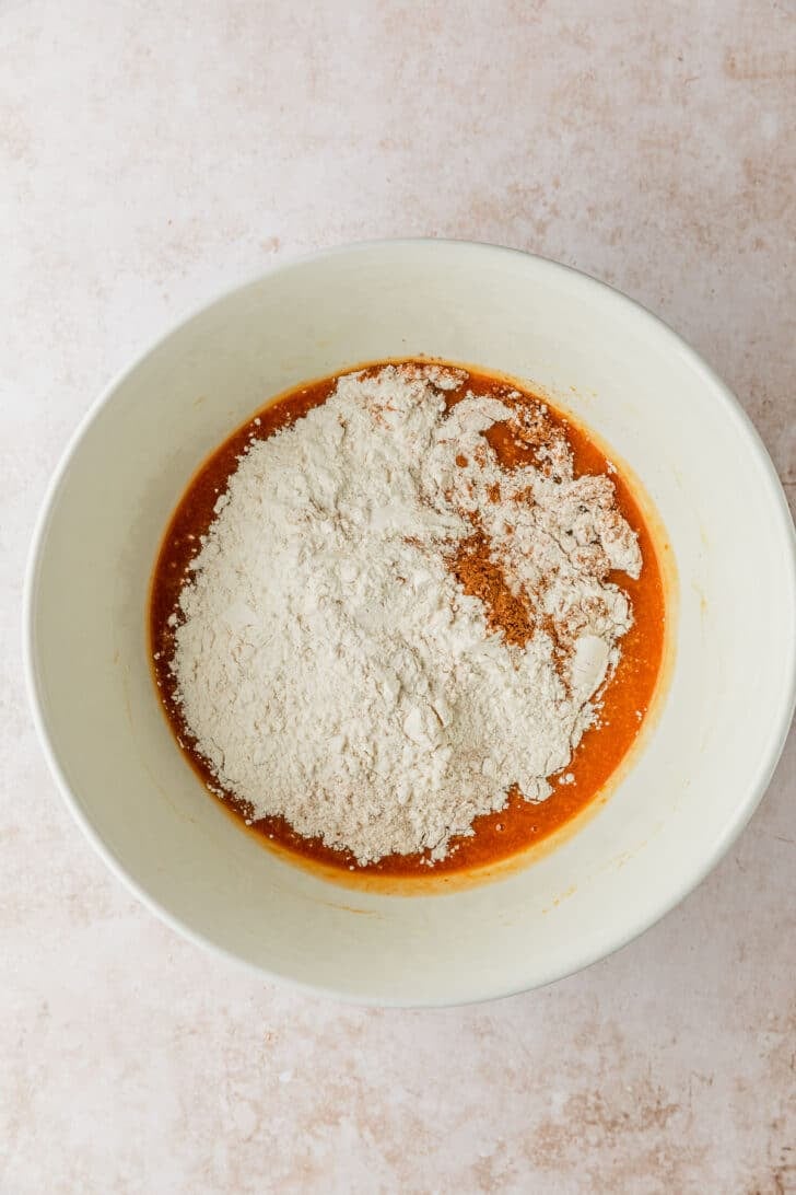 A white bowl of pumpkin mixture topped with flour and spices on a tan counter.