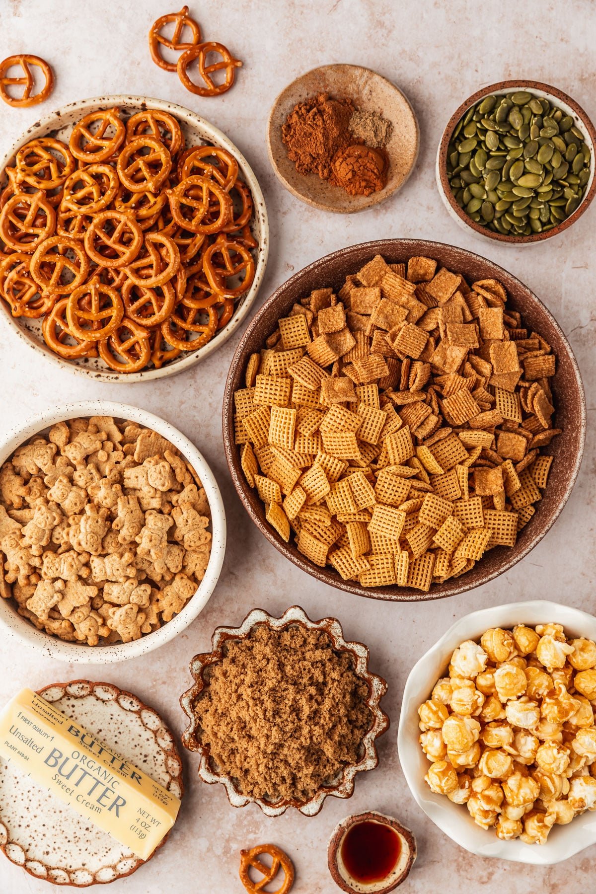 Brown and white bowls of pretzels, spices, pumpkin seeds, cereal, teddy grahams, brown sugar, caramel corn, butter, and vanilla on a tan counter.