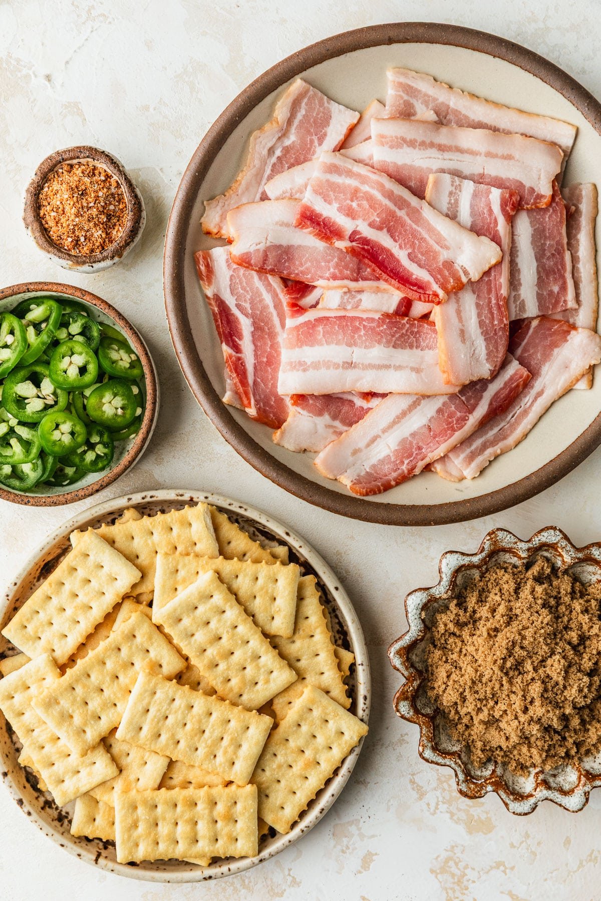 White and brown bowls of bacon, BBQ rub, crackers, brown sugar, and jalapenos on a tan counter.