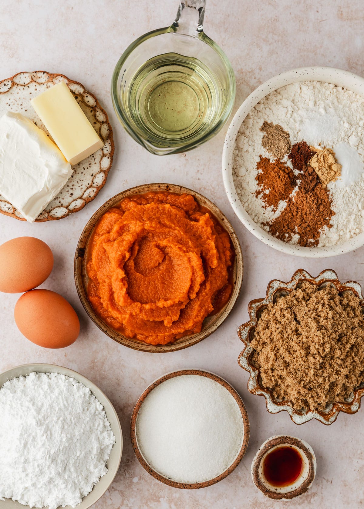 Brown and white bowls of flour, spices, oil, pumpkin puree, cream cheese, butter, brown sugar, white sugar, powdered sugar, and vanilla next to eggs on a tan counter.
