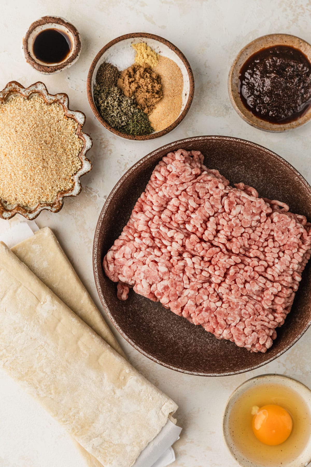 Brown and white bowls of pork, breadcrumbs, spices, apple butter, Worcestershire sauce, an egg, and puff pastry on a tan counter.