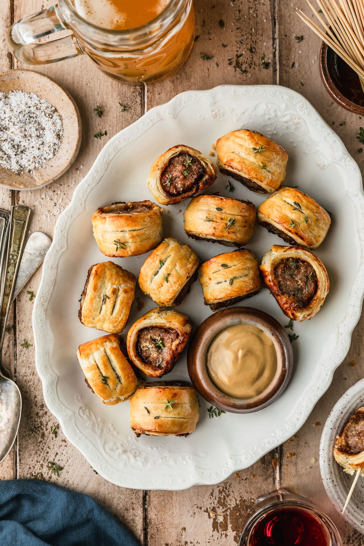 A white vintage platter of pork and apple sausage rolls on a wood table next to glasses of beer and wine, vintage forks, a blue linen, and a brown bowl of toothpicks.
