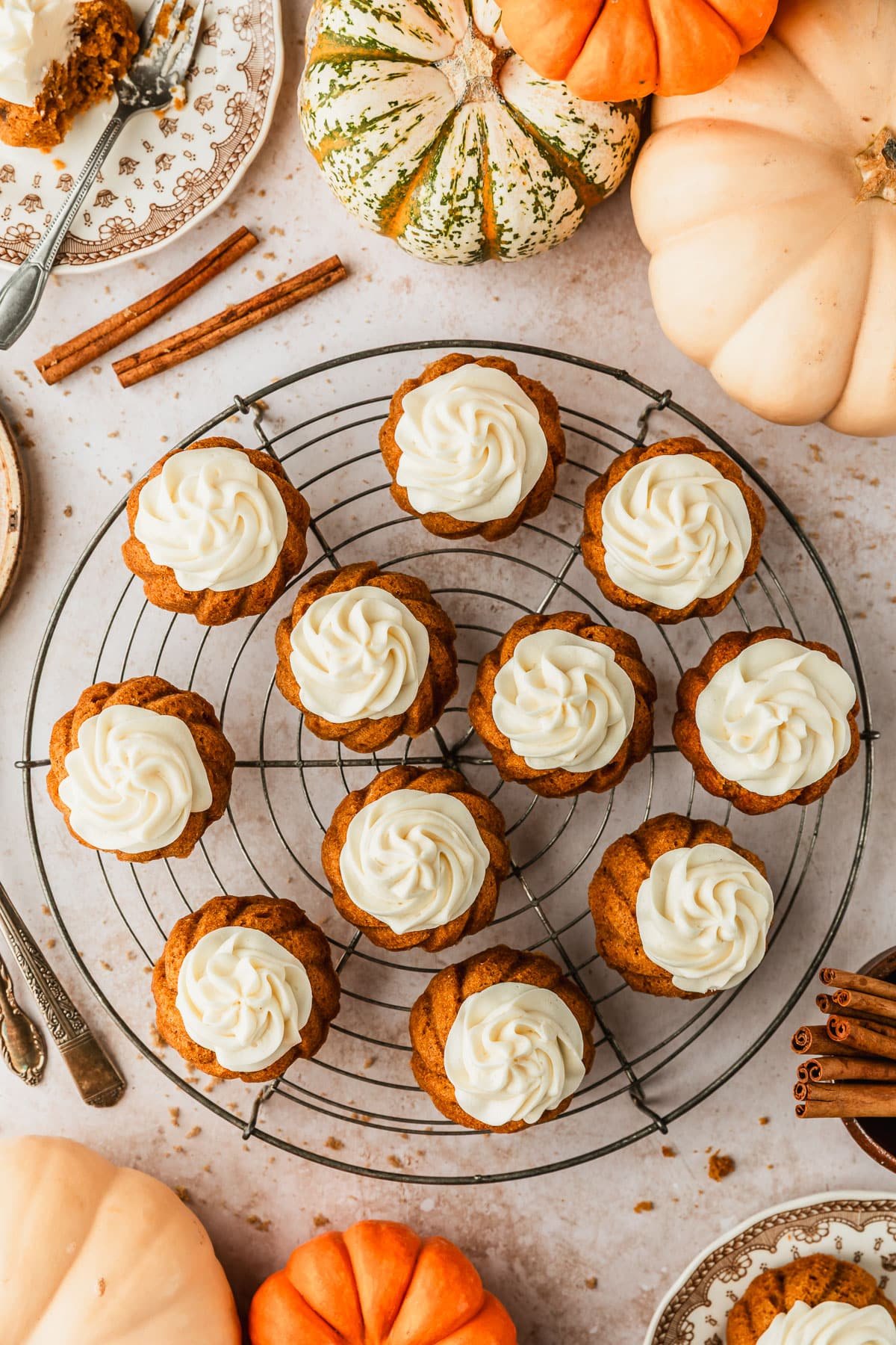 A wire rack of pumpkin mini bundt cakes with cream cheese frosting on a tan marble counter next to brown plates of cake, cinnamon sticks, and pumpkins.