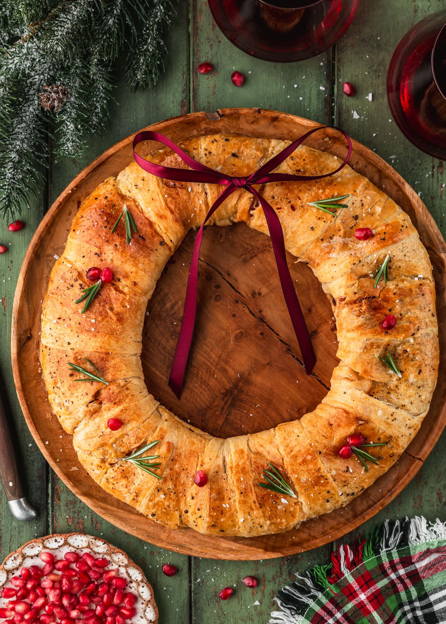 A wood board with a bacon and brie crescent wreath on a green wood table next to garland, glasses of red wine, a plaid linen, and a white plate of pomegranate arils.