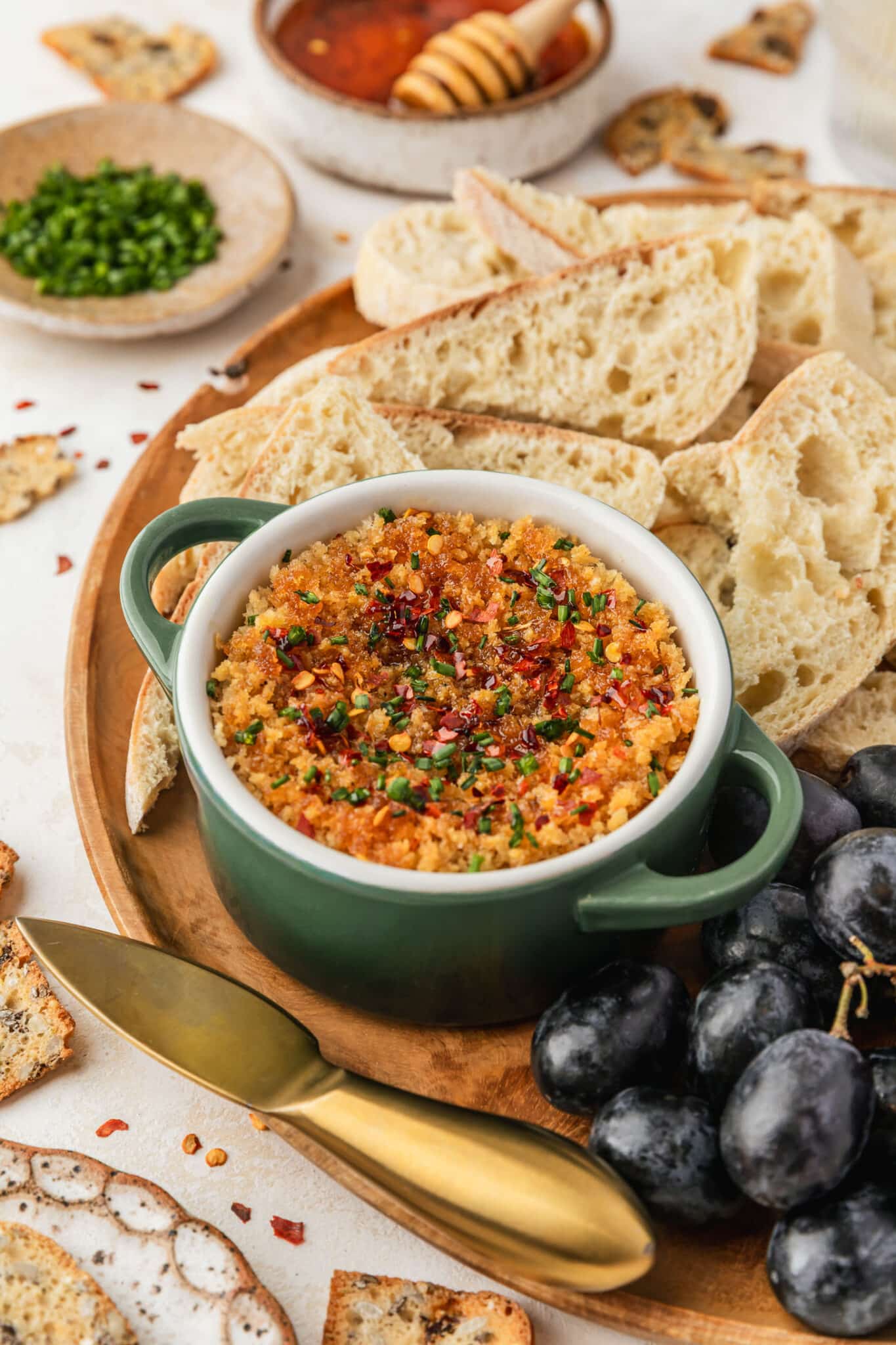 A green ramekin of baked boursin cheese with hot honey on a wood board of bread and grapes next to crackers, glasses of white wine, and brown bowls of honey and chives on a beige counter.