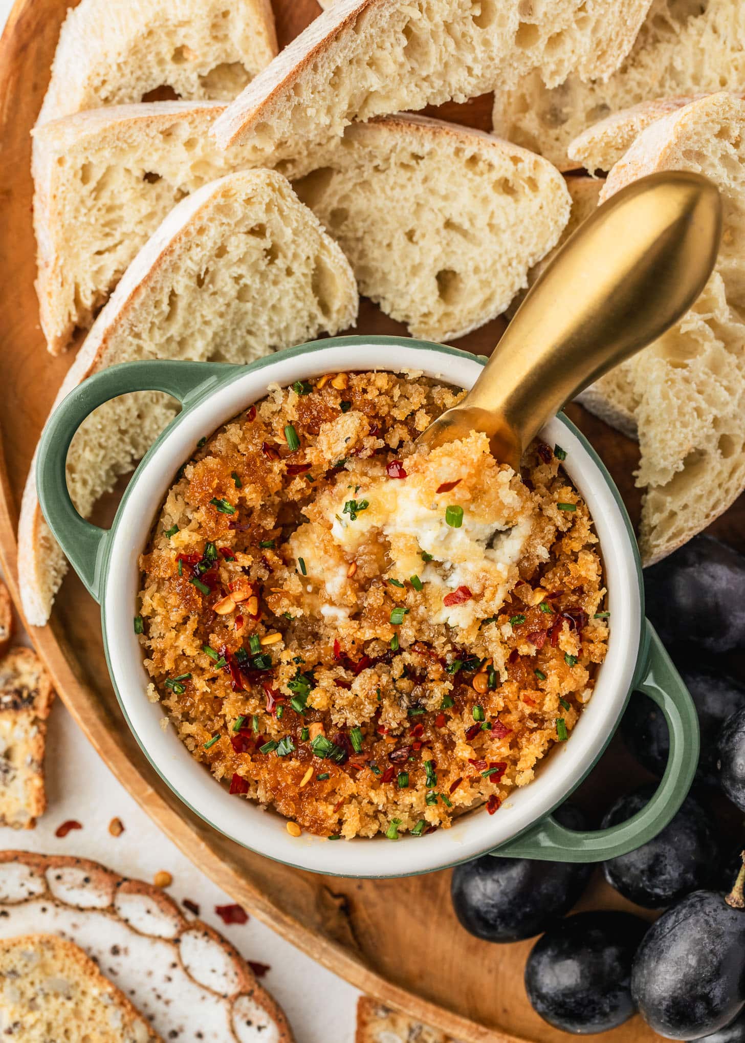 A green ramekin of baked boursin cheese with a gold knife on a wood board next to crusty bread, grapes, and crackers.