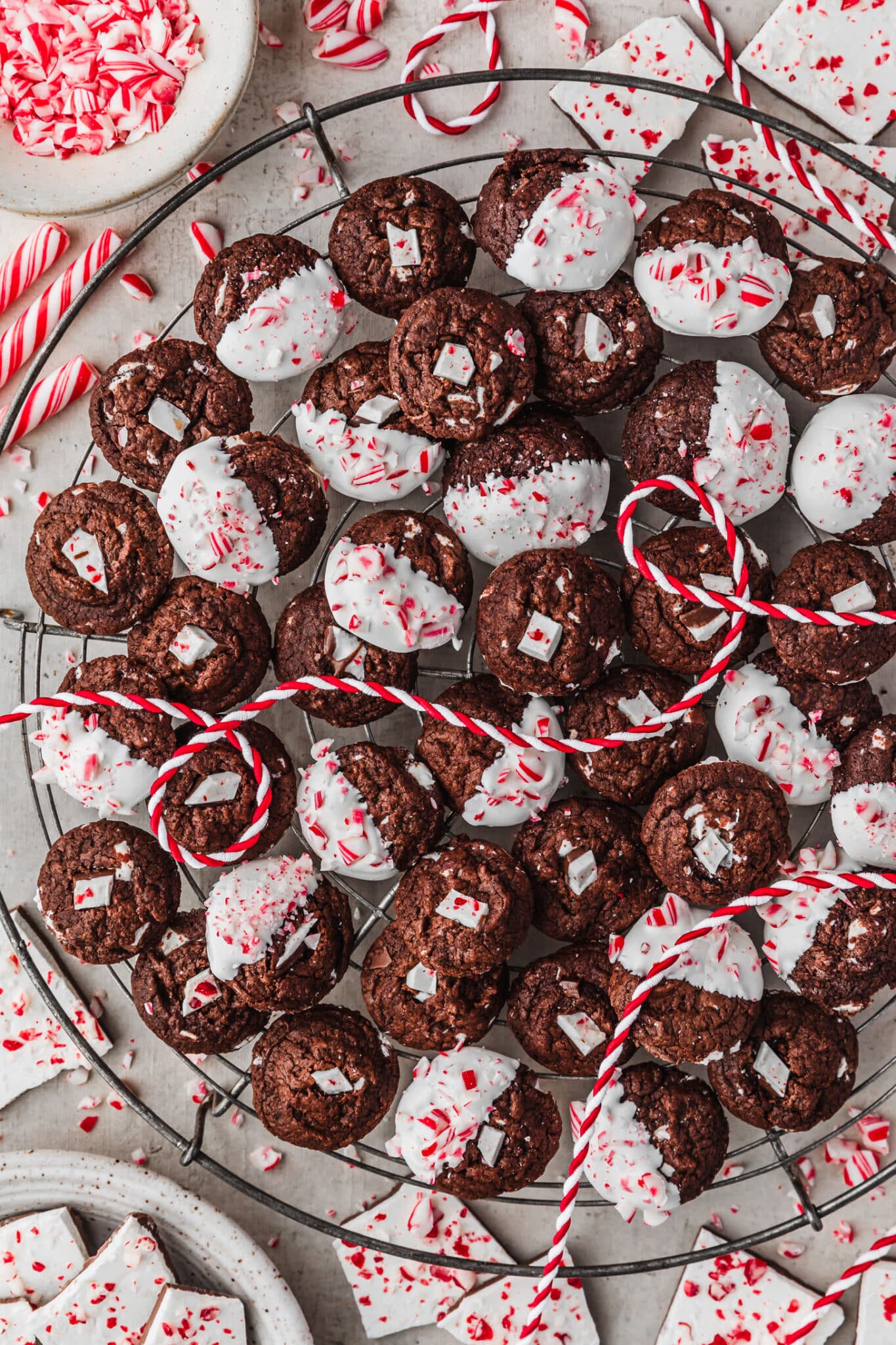 A wire rack of mini chocolate peppermint bark cookies on a grey counter next to a white bowl of chopped candy canes, a white bowl of peppermint bark, and striped red and white ribbon.