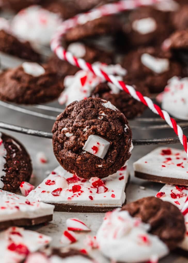 A cookie leaning against a wire rack of mini chocolate peppermint bark cookies next to peppermint bark and red and white striped ribbon with a grey background.