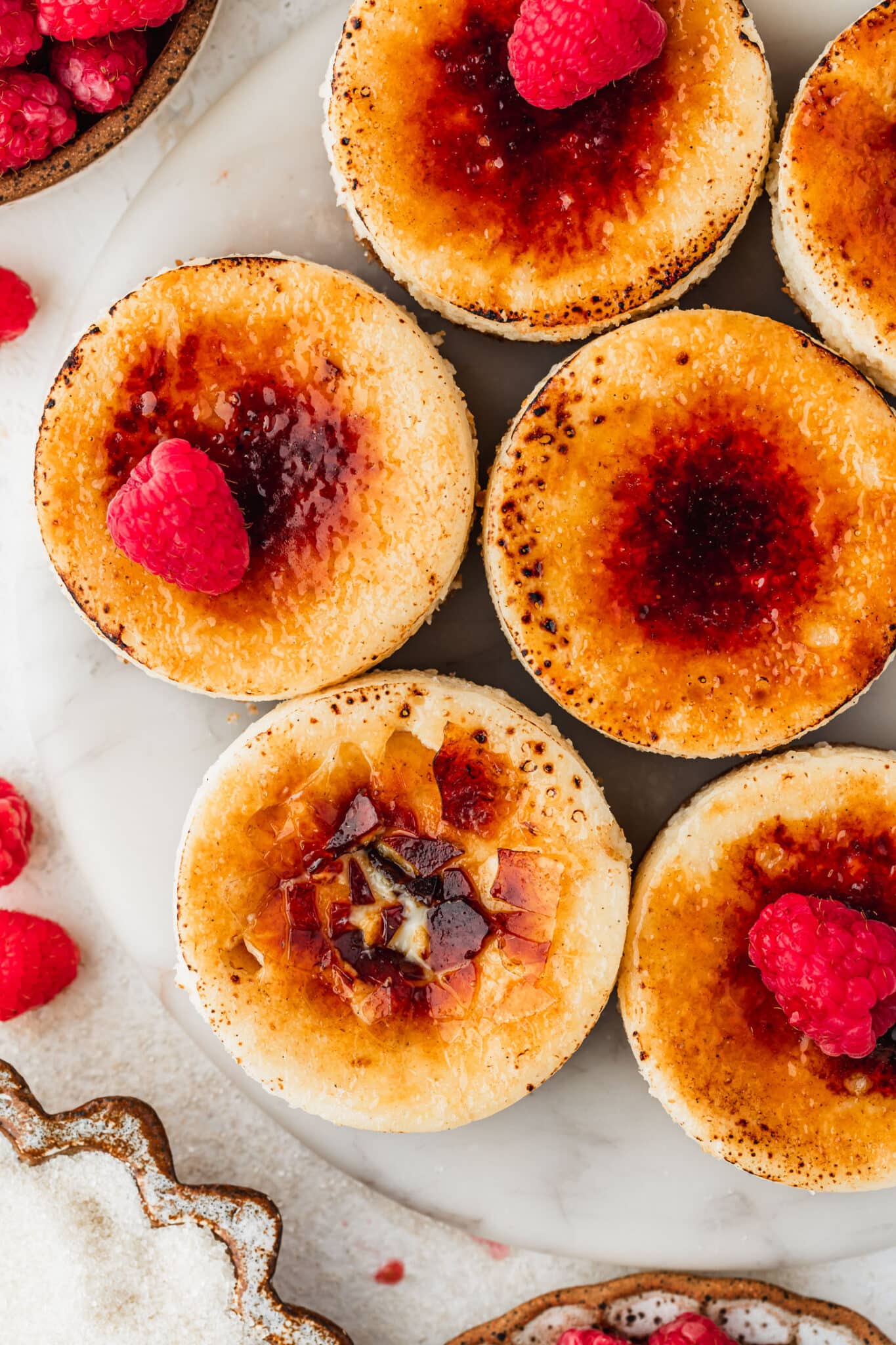 A white platter of mini creme brulee cheesecakes on a white counter next to brown bowls of raspberries and sugar.