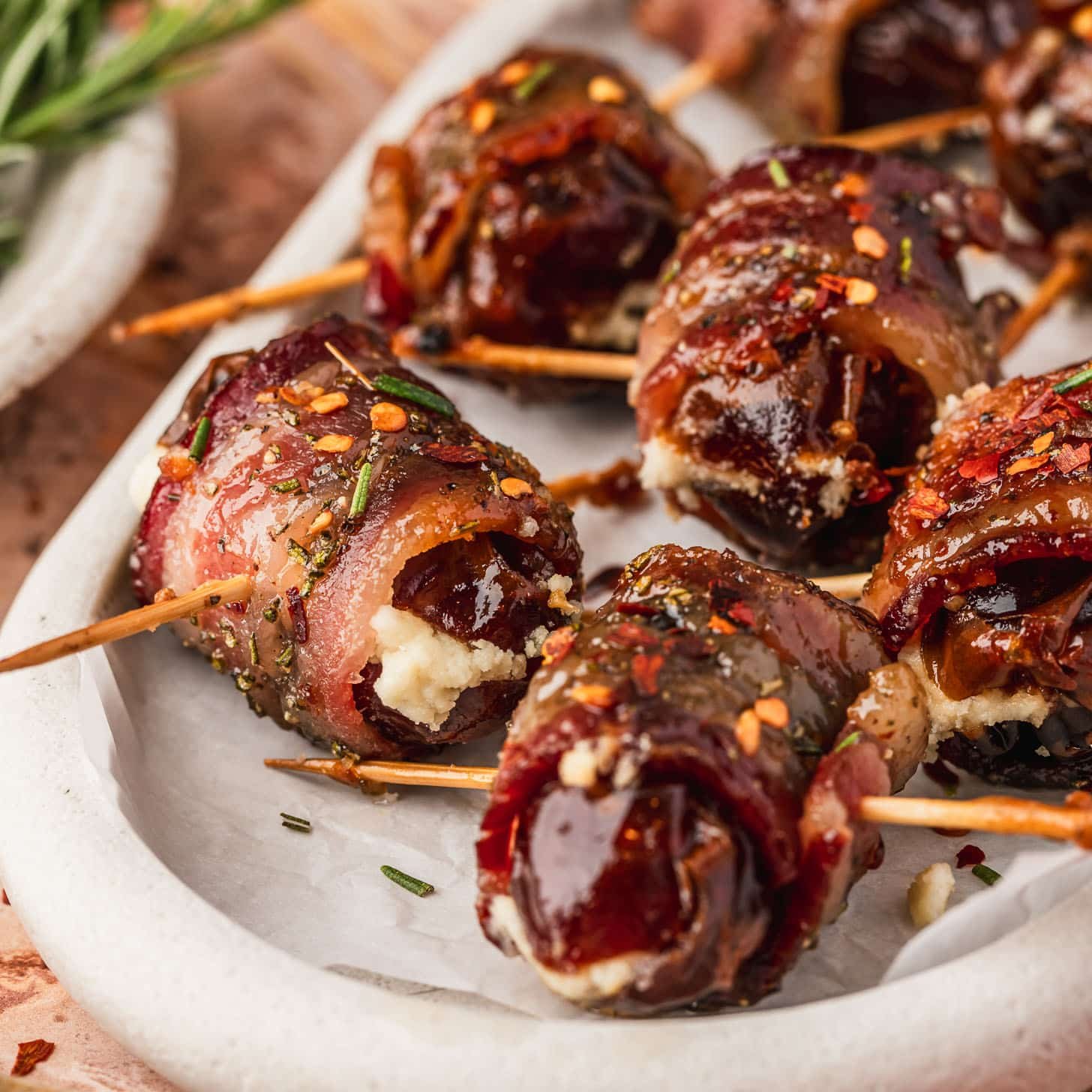 Bacon wrapped dates with goat cheese and maple glaze on a white platter next to glasses of white wine, an orange linen, and a white bowl of rosemary with an orange stone background.