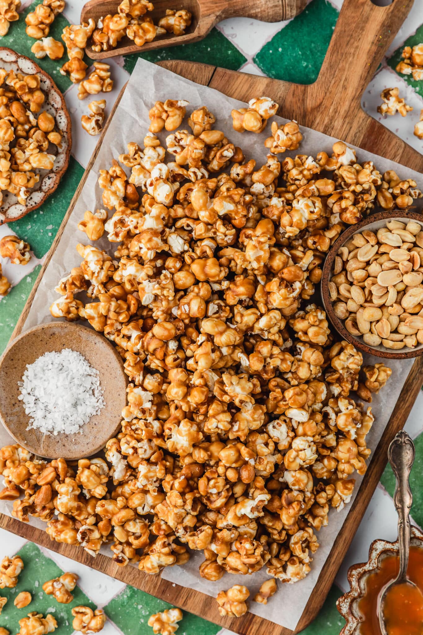 A wood board with salted caramel popcorn and brown bowls of peanuts and salt next to a brown bowl of caramel, a scoop of popcorn, and white bowls of caramel corn on a green and white tiled counter.