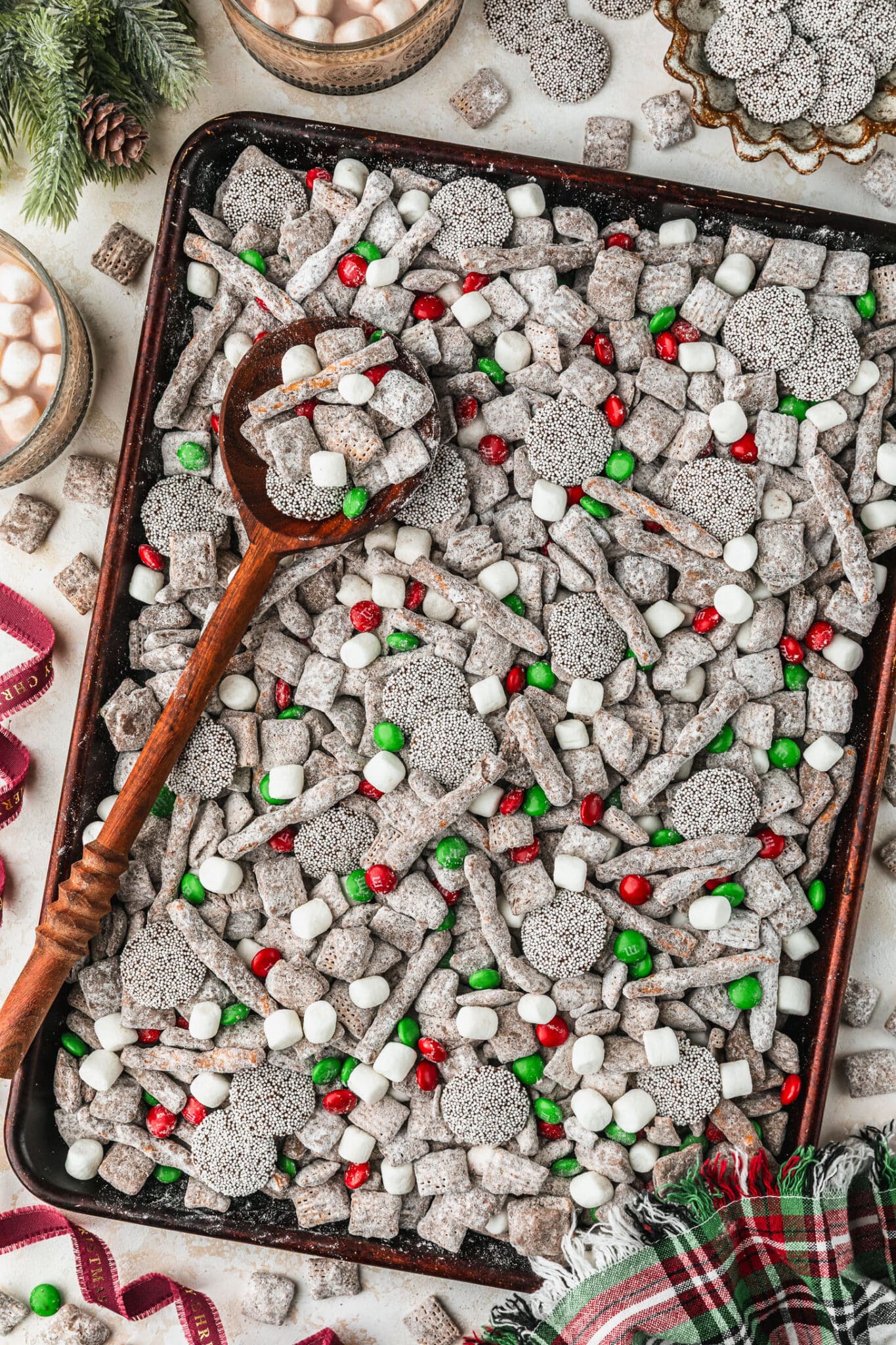 A sheet pan of Christmas muddy buddies with M&Ms and pretzels on a tan counter next to garland, red ribbon, a brown bowl of sprinkle chocolates, and glasses of hot cocoa.