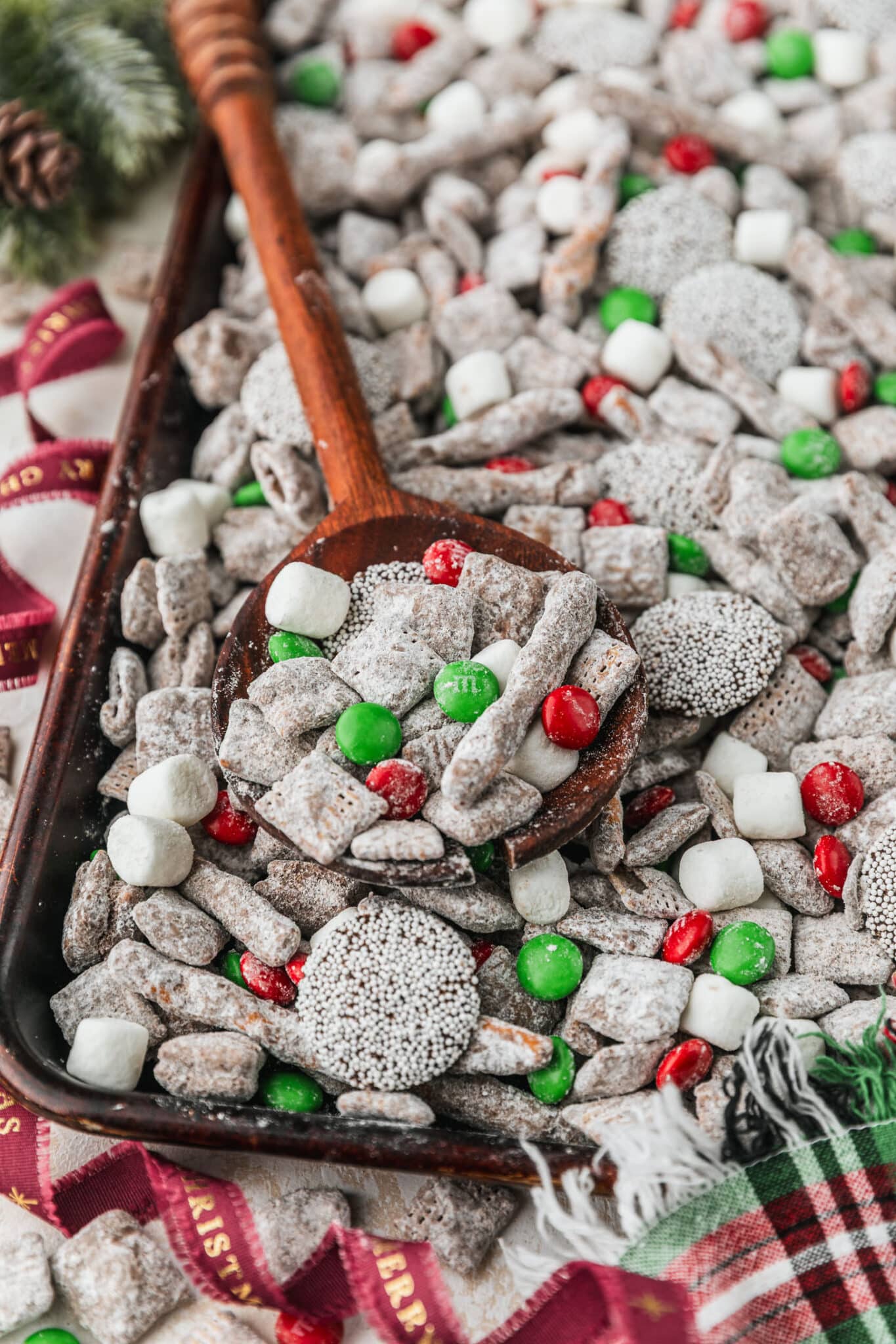 A sheet pan of Christmas puppy chow with a wood spoon on a beige table next to garland, red ribbon, and a plaid linen.