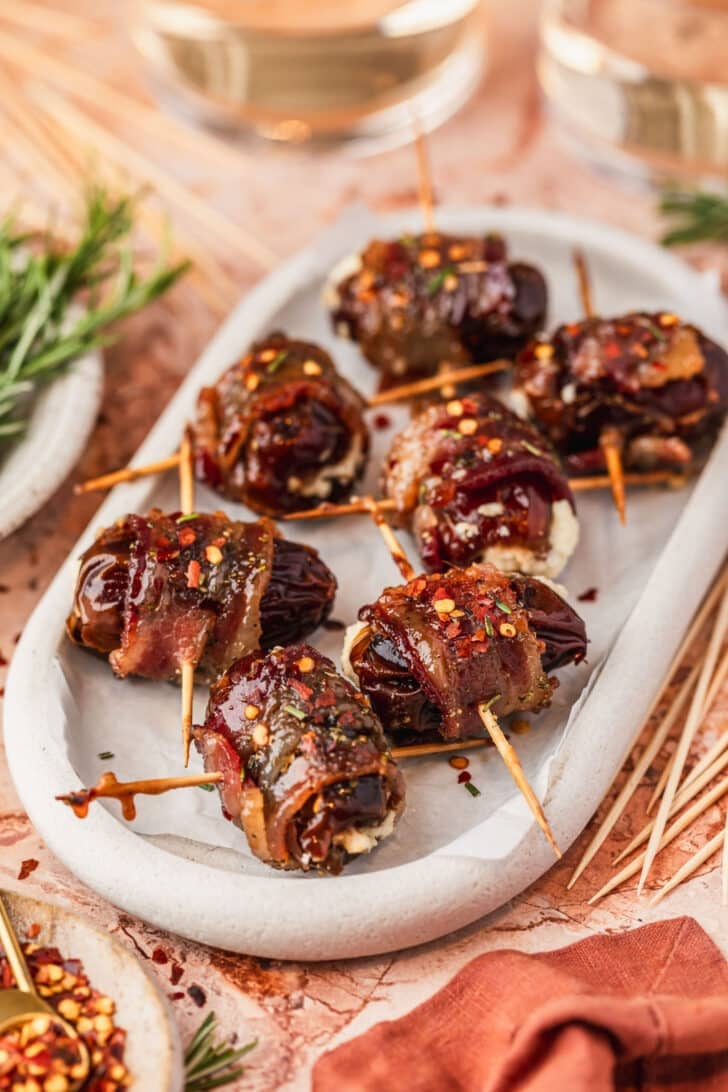 A white platter of bacon wrapped dates with goat cheese on an orange marble counter next to glasses of white wine, a brown bowl of pepper flakes, and an orange linen.