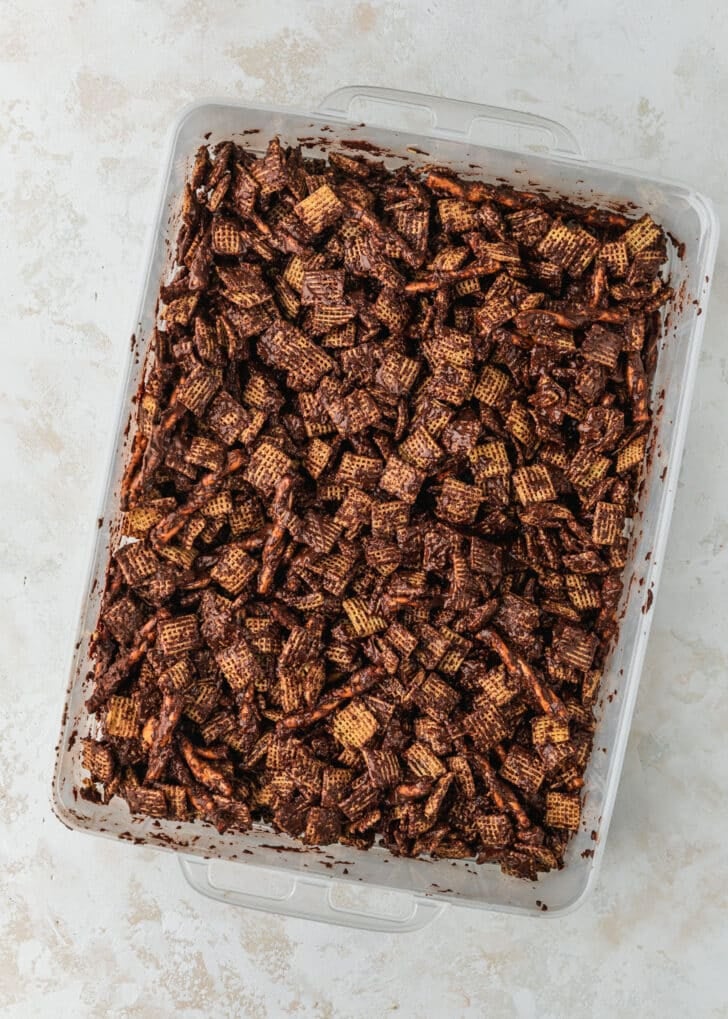 A container of snack mix with chocolate on a tan table.