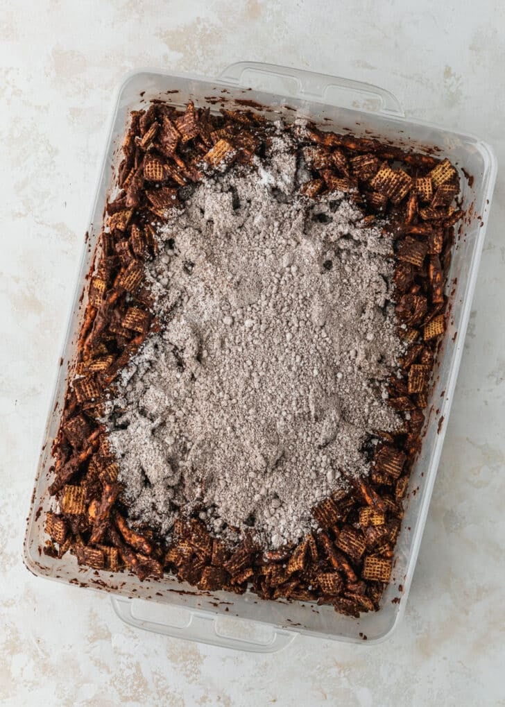 A clear container of hot chocolate Christmas muddy buddies on a tan counter.