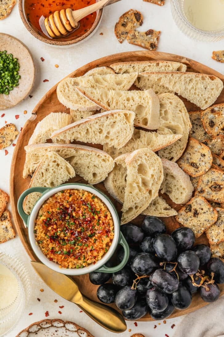 A green ramekin of baked boursin on a wood board of bread and grapes next to crackers, glasses of white wine, and brown bowls of chives and hot honey on a tan counter. 