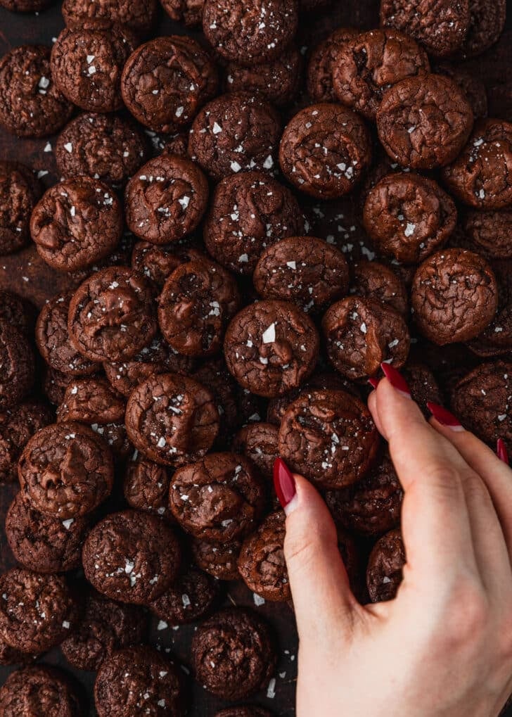 A hand grabbing a cookie from a tray of mini chocolate brownie crinkle cookies.