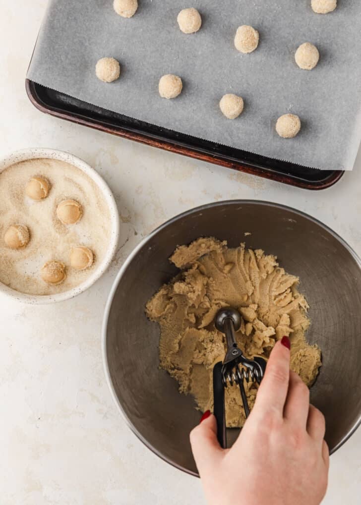 A hand using a scoop to scoop mini sugar cookies dough out of a silver bowl next to a sheet pan of raw cookies and a white bowl of sugar on a tan table.