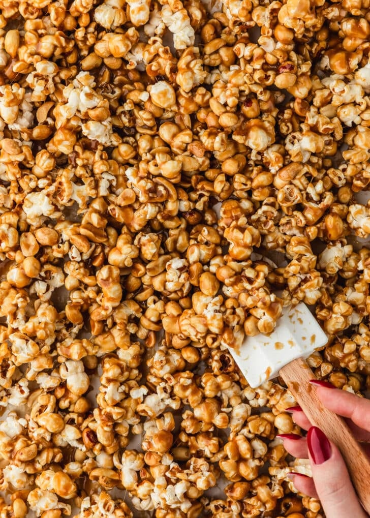 A hand using a rubber spatula to toss popcorn and peanuts with salted caramel sauce.