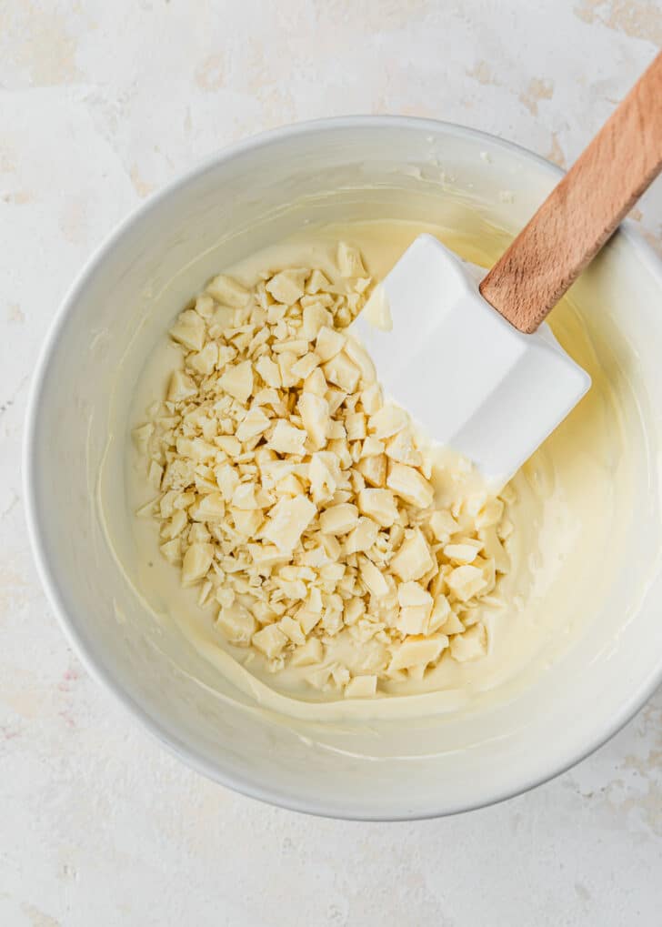 A white bowl of melted white chocolate with chopped chocolate on a tan counter.