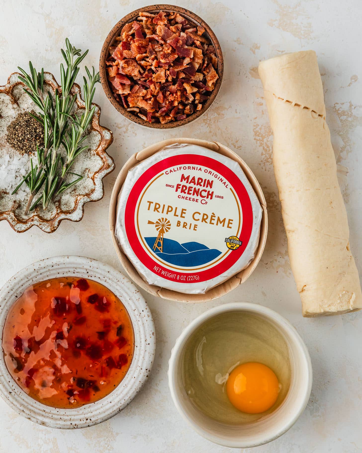 White and brown bowls of bacon, rosemary, an egg, and pepper jelly next to a wheel of cheese and croissant dough on a tan counter.