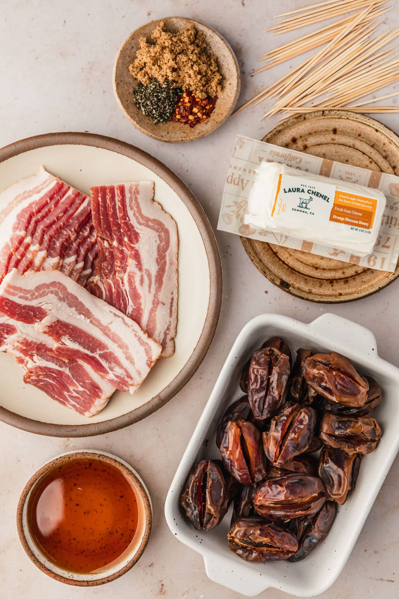 White and brown plates of bacon, dates, goat cheese, brown sugar, rosemary, pepper flakes, and maple syrup on a tan counter.