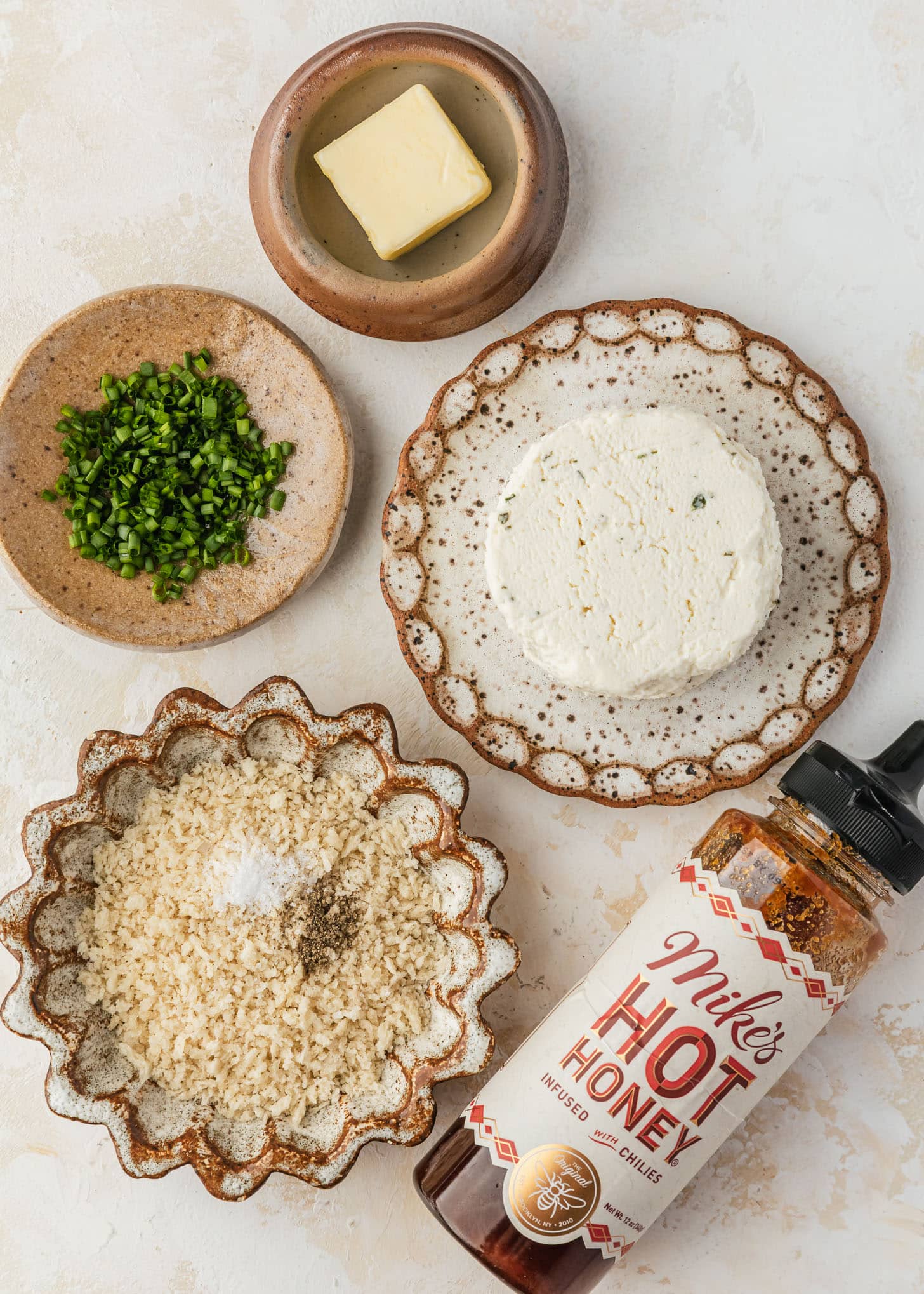 White and brown plates of cheese, chives, butter, and breadcrumbs next to a bottle of hot honey on a tan table.