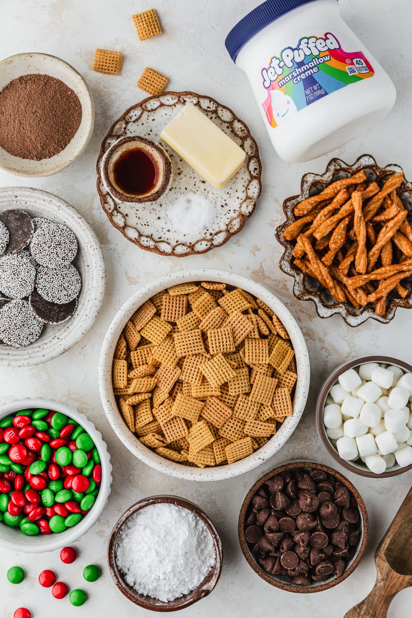 White and brown bowls of hot chocolate mix, pretzels, chex, holiday candies, butter, salt, marshmallows, chocolate, and powdered sugar next to a jar of marshmallow fluff on a tan counter.