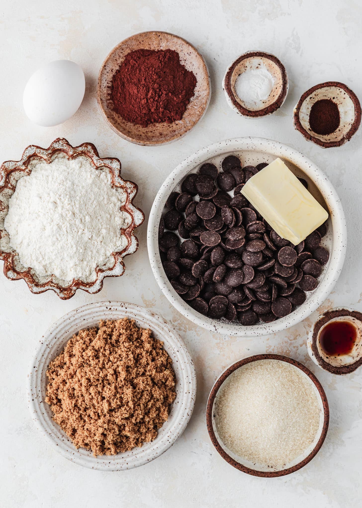 White and brown bowls of chocolate chips, butter, flour, cocoa, espresso powder, salt, baking soda, brown sugar, granulated sugar, and vanilla extract next to an egg on a tan table.
